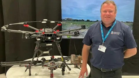 BBC Professor Darren Ansell stands to the right of a table with two drones on it, one large and round in grey and one tiny . He is standing in front of a monitor showing a field under a blue sky and is wearing a navy shirt and black trousers