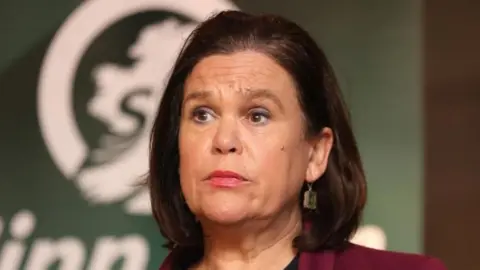 PA Media McDonald is looking to the left of the screen. She has short brown hair and is wearing gold earrings and a reddish blazer.