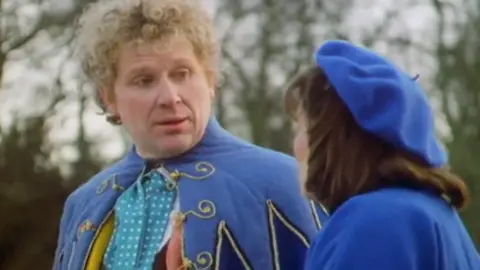 A screengrab from a Doctor Who episode showing the Sixth Doctor, Colin Baker, a man with curly blond hair, wearing a blue robe lined with gold trim. He is talking to a woman with brown hair, wearing a blue coat and a blue beret, whose face is not visible.