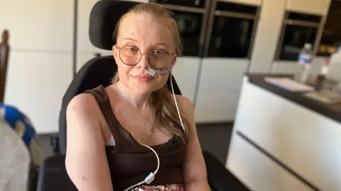 A woman with glasses sits in a wheelchair in a kitchen. She is wearing a colourful skirt, black top and is holding medical equipment.