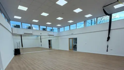 Victoria Goodall An empty dance studio with pine floors, white walls and mirrored wall on the far side.