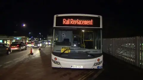 White coach with "Rail Replacement" on the route banner above the windscreen. The photo is taken at night - car headlights can be seen to the left of the bus. There is a fence to the right of the bus. There are road cones between the bus and the traffic
