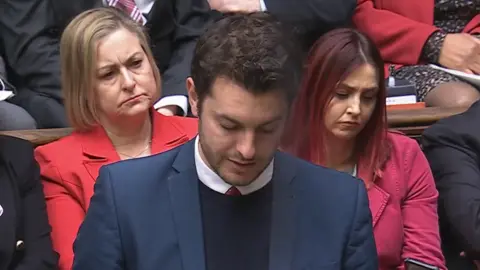 Parliament Live Jack Abbott speaks in the House of Commons during Prime Minister's Questions. He looks down at notes he holds in his hands while he talks. MPs behind him look on. Abbott has short dark hair and wears a navy suit with a black jumper underneath, white shirt and red tie.