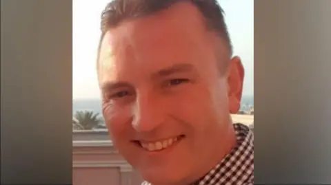 A man wearing a chequered shirt smiles while looking into the camera.