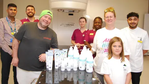 A dark glass table is at the centre of the picture. On top of the table are white and blue pots, tubs and bottles of skin care products. Seven adults and one child are standing around the table looking at the camera. Some are wearing hospital uniform, three are wearing white t-shirts. They appear to be in a radiotherapy room with a large piece of medical equipment behind them. 