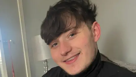 Lisa Petrie John Mcnab smiles at the camera while sitting on a couch in a living room. He has short dark brown hair and wears a dark top.