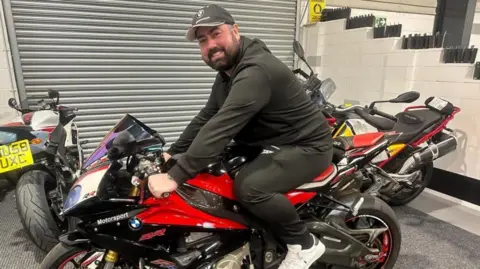 A man wearing a black hoodie and a cap smiles as he straddles a large red and black motorbike in a garage.