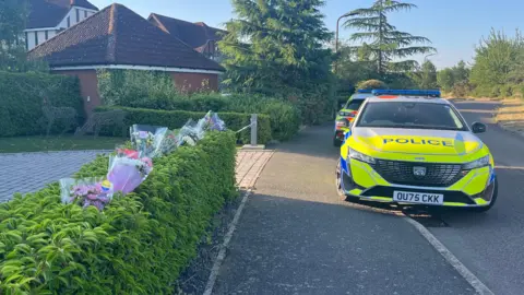 Holly Nichols/BBC The scene at Carnoustie Drive in Great Denham. On the left is a hedge which has plastic-wrapped flowers sticking into it, and beyond is an entrance to a driveway with police tape across it. On the right are two police cars parked partly on the pavement. Beyond it is another detached house and trees.