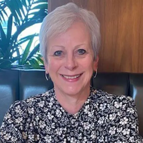 Judith Logan Judith Logan is smiling broadly. She has short grey hair and she is wearing a black, white and grey floral blouse. She has hooped earrings and is sitting on leather seating. Judith Logan understands life-saving surgery must take priority


