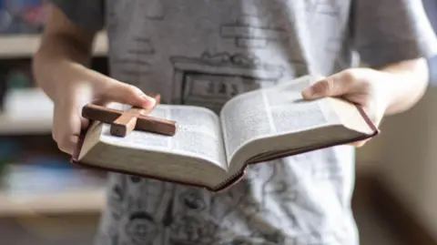 Someone holds a book in their hands with brown edging, while holding a brown cross in their right hand.