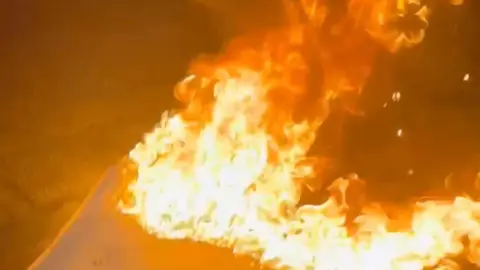 Flames engulf a mattress by the side of the road.