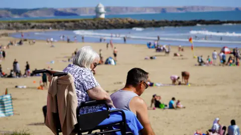 Getty Images A woman with white hair sits in her wheelchair looking over the crowded beach on a glorious hot sunny summer's day. A man sits besides her on the path.
