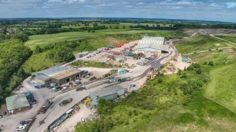 An aerial view of the quarry, surrounding by green fields. Numerous buildings separated by roads and car parks can be seen.