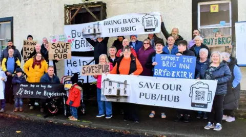 Members of the Brydekirk community gathered in front of the former pub holding an array of banners titled with slogans such as 'save our pub/hub'.