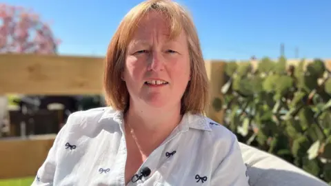 Sharon Barbour/BBC Nadeen is sitting in a sunny garden wearing a white blouse. A microphone is attached to her top. A hedge and fence can be seen in the background which is blurred. 