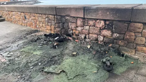 BBC The aftermath of a fire on a paved walkway near a stone seawall. The ground is covered in melted green plastic material with charred debris and remnants of a green wheelie bin. The fire has also left black scorch marks on the stone wall behind it. The ocean and a distant shoreline visible in the background.