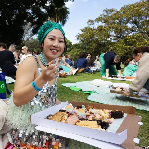 Jessica Hromas/BBC A woman wearing a green headband and a sequined dress, eating cake from a large pizza box with several different slices of cake