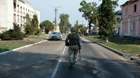 REX/Shutterstock A Ukrainian serviceman walks near damaged buildings in the city center of Sudzha, in Ukraine-controlled territory of Russia's Kursk region in August 2024