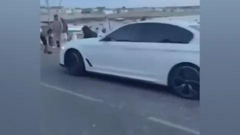 A screen grab from a social media video of a white car and people standing around it