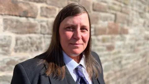 Kerry Tough has shoulder-length, brown hair. She is wearing a suit jacket, waistcoat, shirt and tie. There is a stone wall behind her