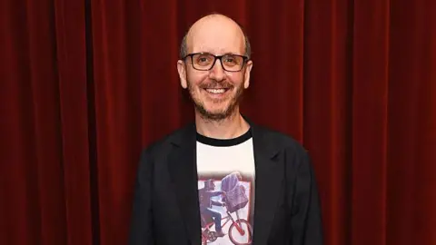 Dave Benett/Getty Images Jack Thorne stands in front of a burgundy curtain, wearing an E.T. Top and a black blazer.