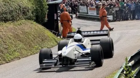 A white formula 1 style racing car on a tarmac track bordered by a grassy verge and embankment. Crowds stand behind a railing at the side of the track and lean over barriers at the side while crew in orange jumpsuits walk across it behind the vehicle.