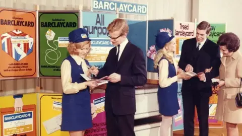 Barclaycard A retro photo from the 1960s showing Barclaycard staff promoting the credit card with a display stand behind them. The female staff wear cream blouses under blue short pinafore dresses and a branded blue cap. One speaks to a smartly dressed man and woman, while the other talks to a man in a suit. 