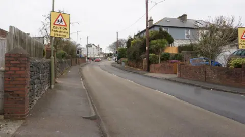 Marldon Road in Paignton. Yellow signs saying "CAUTION Pedestrians crossing" are on signposts. It is an overcast day.