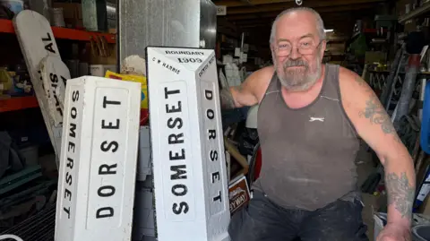 Two white signs on the floor, standing up, saying "Dorset" and "Somerset". There is a man with a beard crouching down next to them.
