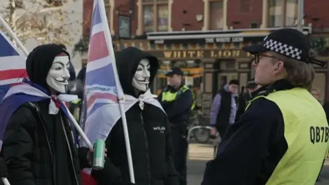 Three men wearing face masks in the style of the film V for Vendetta, white masks with black facial features, dressed in black and carrying Union flags, looking at two police officers facing them