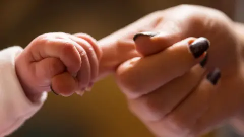 A close-up of a baby's hand grasping an adult's index finger. The adult has dark, painted nails.