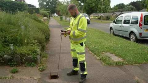 A member of staff for South West Water - wearing high viz clothing - stands in a residential street. He is holding a long metal pole that sits in the access hole of a water pipe. He is monitoring data on his mobile phone. 