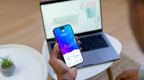 Trader's hand holding a phone showing financial information with a laptop on a similar page behind.