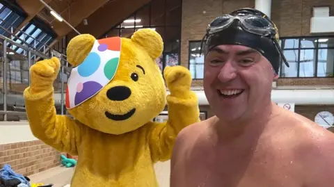 BBC Pudsey, a human-sized yellow bear mascot wearing a patch over his eye, stands with his arms in the air. Next to him is Edward Sault, a topless man wearing goggles and a swimming cap.