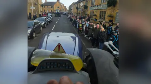 Somerset Tractor Runners A view from the cab of a tractor. A crowd of people has gathered outside a pub to watch and wave at the line of tractors coming up the street.