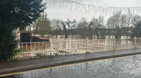 The image shows lots of water both on a road and what looks to be fields. The water on the fields is brown and seems to have risen high up the trees in the background.