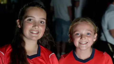 Jamie Niblock/BBC Amelia, who has long brown hair, and Imogen, who is blonde, are both smiling while wearing the red England away top.