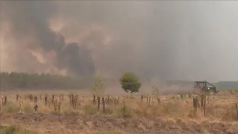 A tractor on the right hand side of the frame. Thick smoke fills the sky above farmland and forestry