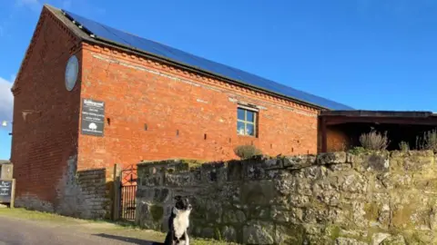 Babbinswood Farm Community Benefit Society A large red brick building with a stone wall running alongside it and a black and white dog sat in front