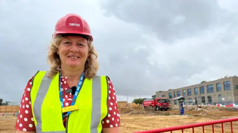 Woman with hard hat and high-vis vest stands infront of a metal fence. The fence is sectioning an area which has been demolished. A building is in the background.