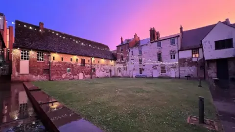 Gloucester History Festival Blackfriar's Priory is in the background of its grass garden. There are fairy lights twinkling and a blue, purple and orange sunset sky behind.