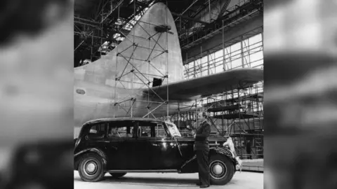 A black and white photo of a BBC reporter in front of the partially-completed Brabazon inside a large aircraft hangar