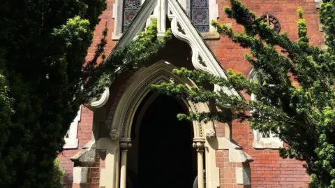Local Democracy Reporting Service The main entrance to a Grade II listed church, showing the white stone archway and gabled roof, surrounded by the rest of the red brick building.