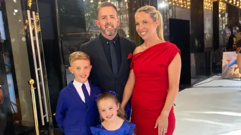Tom Smith A blonde woman in a red dress, man in a black suit, and two children in royal blue smile at the camera. The background is plain but looks expensive - they are in the Raffles hotel in london.