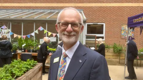 Jessica Bayley/BBC An older man with short white hair and beard is smiling at the camera. He has black round glasses and is wearing a suit jacket with a white shirt and floral tie. He is standing in front of flower beds and a table with potted plants on it. In the background there is a greenhouse with bunting hanging from it.