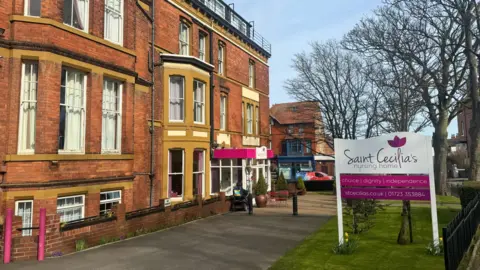 BBC/Emily Johnson A tall red-brick building with a pink reception entrance. A white and pink sign at the front reads "Saint Cecilia's nursing home".