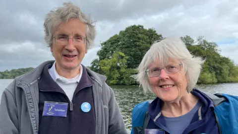 Edward and Amanda Bevan are standing in front of a lake. Edward has grey hair and wears glasses. He is wearing a grey zip jacket and purple jumper. He has a "volunteer" badge with a picture of a heron on it. Amanda has a grey bob and wears glasses. She is wearing a blue jacker with a purple jumper, and also has a heron volunteer badge.