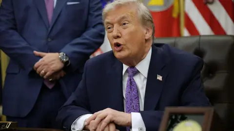 Donald Trump at a desk in the oval office. He is wearing a navy suit. 