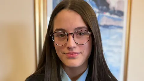 Pippa smiles in her school uniform. She has long brown hair and glasses. On her navy blazer there is a red poppy pin.