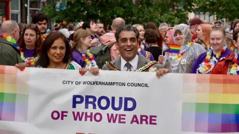 A gathering of people with colourful clothes and colourful flags behind a large flag which reads city of wolverhampton council, proud of who we are